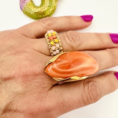 Anel largo dourado com pedras coloridas numa mão com unhas roxas e fundo branco.