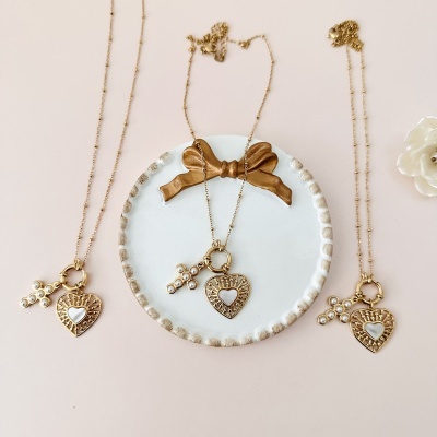 Três colares dourados com pendentes em forma de coração e chave com pérolas sobre fundo claro.