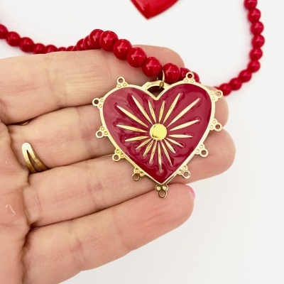 Colar de contas vermelhas com pendente em coração vermelho e detalhes dourados, mostrando uma mão a segurá-lo.