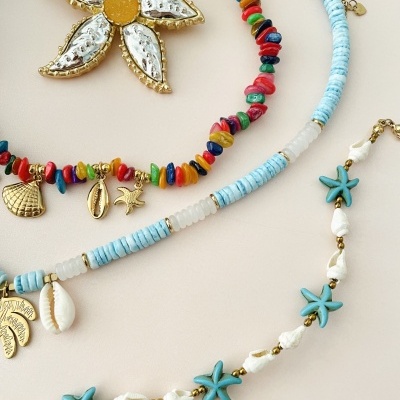 Três colares com pedras coloridas, conchas e pingentes dourados sobre fundo bege.