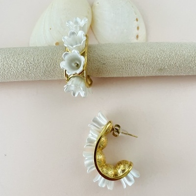Brincos dourados com flores brancas, sobre fundo claro e conchas brancas ao fundo