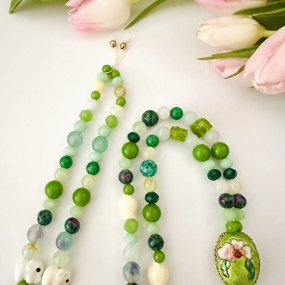 Colar colorido com contas verdes e pingente oval com flores, sobre fundo branco com tulipas rosa.