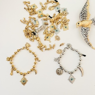 Pulseiras douradas e prateadas com pendentes variados e broche de pássaro em fundo branco