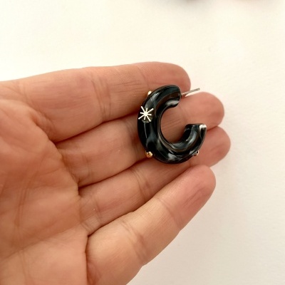 brinco argola preta pequena com detalhes dourados e estrela, segurado numa mão