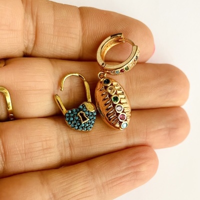 Brincos dourados com pedras coloridas, incluindo coração, oval e argola, sobre dedos de mão