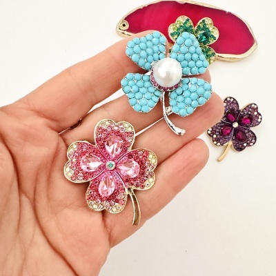 Broches decorativos em forma de trevo coloridos com pedras, sendo azul, rosa e roxo, mostrados numa mão.