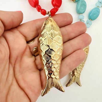Colar com contas vermelhas e pingente em forma de peixe dourado, e colar azul-turquesa com pingente peixe, mão a segurar pingente dourado