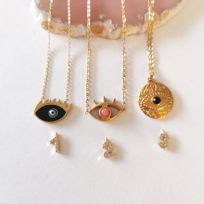 Três colares dourados com pendentes de olhos e medalhão, exibidos em suporte de pedra rosa e branca, cada um numerado de 1 a 3.