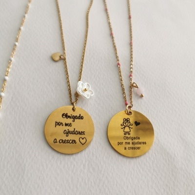 Dois colares dourados com pendentes redondos e inscrições em português, colocados sobre uma superfície clara.