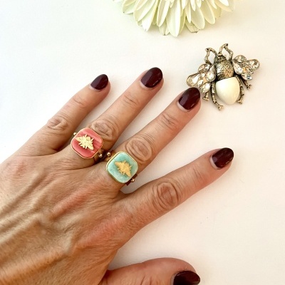 Anel dourado com pedras rosa e azul e broche de abelha em superfície branca