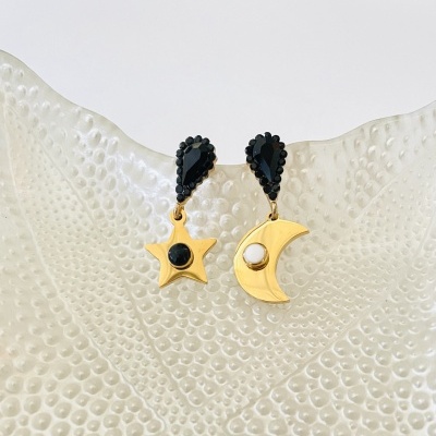 Brincos dourados em forma de estrela e lua com detalhes pretos e branco sobre superfície branca texturada.