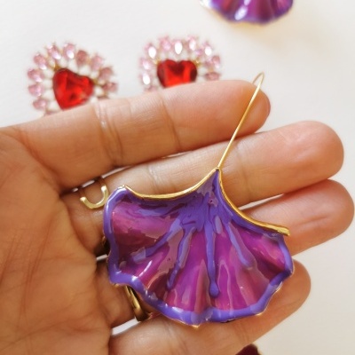 Brinco esmaltado roxo com borda dourada em forma de folha na mão, com outros brincos ao fundo