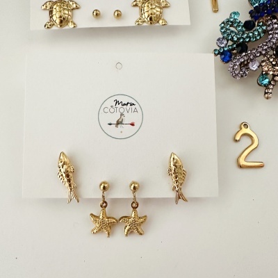 Conjunto de brincos dourados com tartarugas, peixe e estrelas-do-mar em cartão branco com logotipo Maria COTOVIA, e pendente dourado número 2.