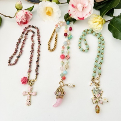 Três colares com pendentes decorativos sobre fundo branco com flores coloridas ao fundo.