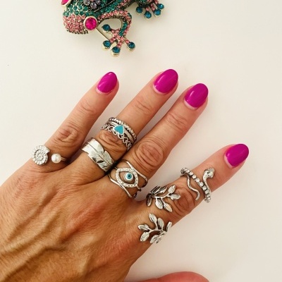 Mão com ungüentos rosa e vários anéis prateados, com pulseira de rã colorida ao fundo