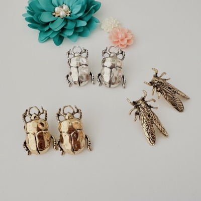 Brincos metálicos em forma de insetos com flores coloridas ao fundo