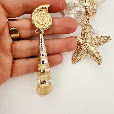 Brincos dourados com design de concha e estrela do mar, um segurado por mão sobre fundo branco.