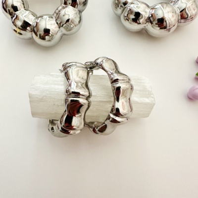 Brincos grandes prateados com design arredondado em seções, sobre superfície branca e luz translúcida.