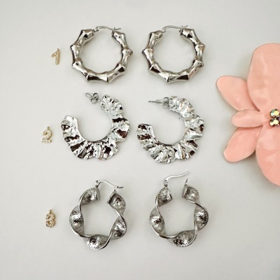 Três pares de brincos prateados com diferentes texturas e formatos sobre fundo branco, ao lado uma flor rosa