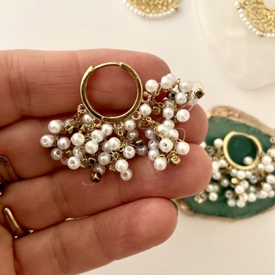 Brinco dourado com pérolas e cristais castanhos segurado por uma mão, outros brincos ao fundo.