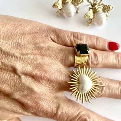 Anel dourado com pedra preta e anel dourado com design de sol com pérola em forma de coração numa mão com unhas vermelhas, brincos de abelhas douradas com pérolas no fundo
