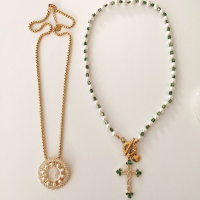 Dois colares, um dourado com pendente redondo de pérolas e outro com contas brancas e verdes e cruz dourada com detalhes verdes.