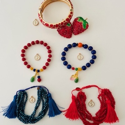 Acessórios incluindo pulseira dourada com padrão vermelho, brincos de morango, pulseiras de contas vermelhas e azuis com penduricalhos, pulseiras com franjas azul e vermelha