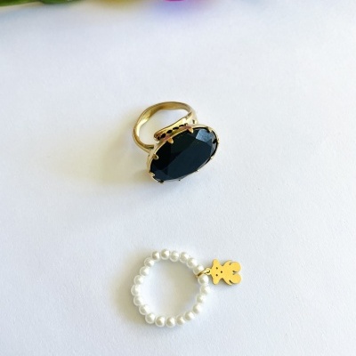 Dois anéis, um dourado com pedra preta e outro de pérolas com pendente dourado, sobre fundo branco com flores ao fundo.