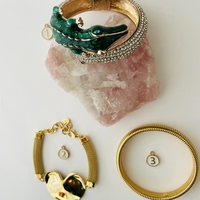 Três pulseiras douradas, uma com crocodilo verde e pedras, sobre pedra rosa, outras duas douradas com etiquetas numeradas