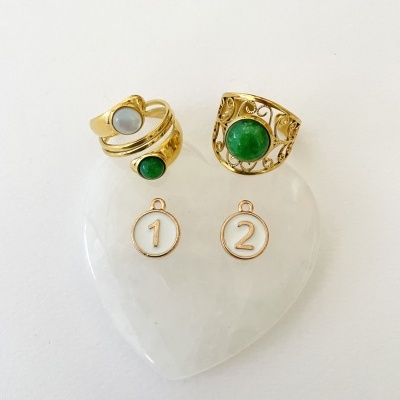 Anéis dourados com pedras verdes e cinzentas sobre coração branco translúcido com pendentes '1' e '2'