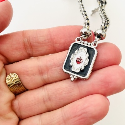 Colar com pendente rectangular preto e prata com símbolo vermelho, com corrente bicolor e pingentes, numa mão.