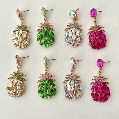 Brincos em forma de abacaxi com cristais coloridos em dourado, verde, branco iridescente e rosa, dispostos em duas filas sobre fundo branco.