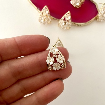 Brinco trianglar com flores brancas e detalhes dourados segurado por mão e base vermelha ao fundo