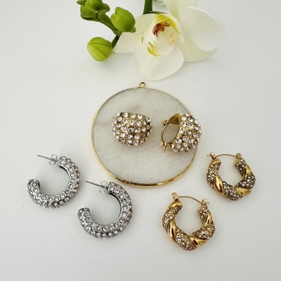 Três pares de brincos de argola com pedras brilhantes em metal dourado e prateado junto a flor branca