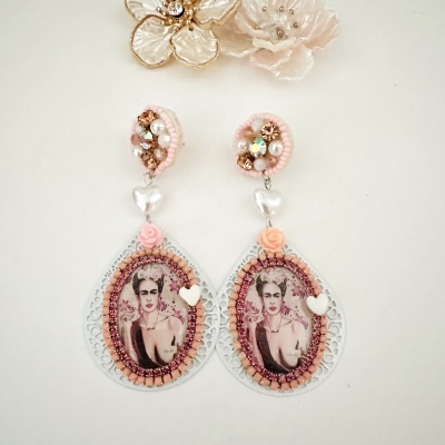 Brincos grandes com imagem da pintora Frida Kahlo e decoração em pérolas e rosas em fundo branco.