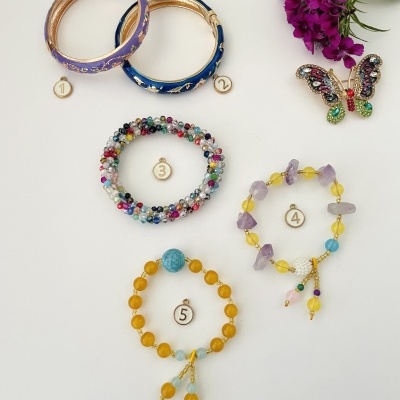 Cinco pulseiras variadas numeradas em fundo branco ao lado de broche de borboleta e flores roxas