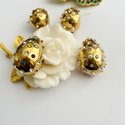 Conjunto de bijuteria dourada com pedras coloridas, flor branca de porcelana e broche colorido ao fundo