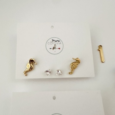 Conjunto de brincos com cavalo-marinho dourado e pedras brilhantes em cartolina branca com logo Marta COTOVIA