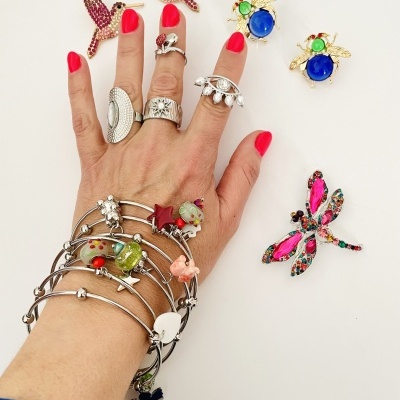 Mão feminina com vários anéis e pulseiras coloridas e brincos de pássaros e libélula sobre fundo branco