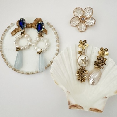Brincos com pedras azuis, pérolas, pendentes e decoração floral dispostos em prato e concha branca.