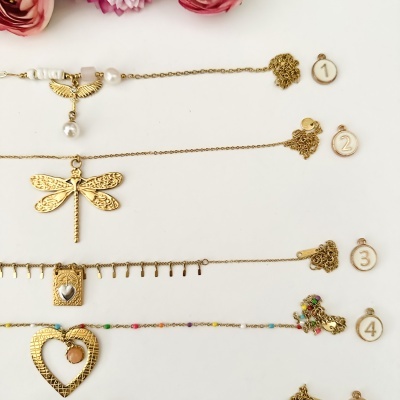 Cinco pulseiras douradas com pendentes variados e contas coloridas sobre fundo branco e flores rosas