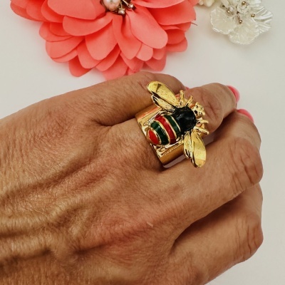 Anel dourado com abelha colorida numa mão com flores decorativas ao fundo