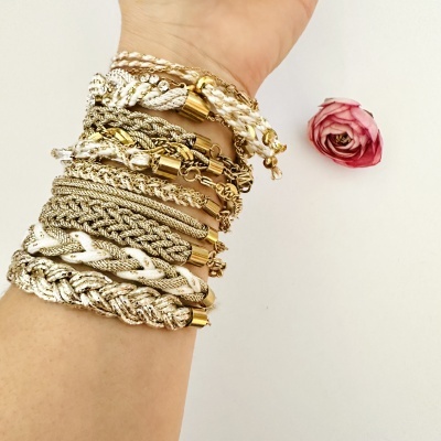 Pulseira artesanal trançada com fios beige, branco e dourado no pulso, com uma flor rosa ao lado.