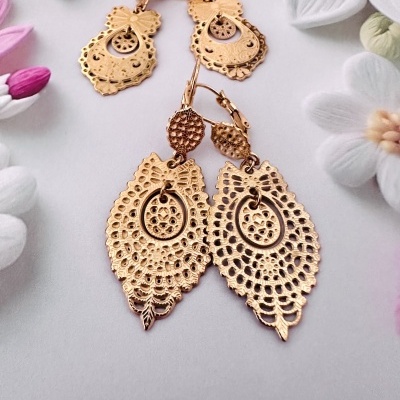 Dois pares de brincos dourados com padrão rendilhado sobre fundo cinza com flores artificiais