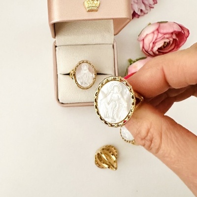 Anel dourado com camafeu branco dentro de caixa rosa, decorada com coroa dourada, com flores rosas ao fundo