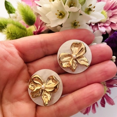 Brincos redondos brancos com flor dourada em relevo no centro, sobre mão, fundo com flores coloridas.