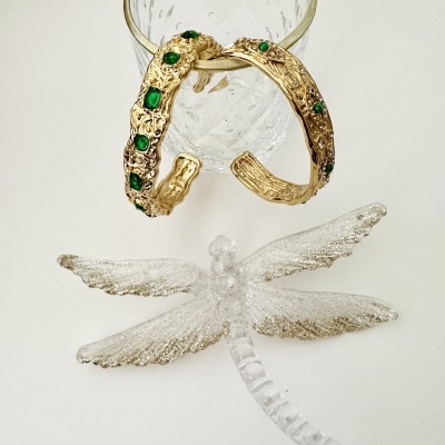 Par de pulseiras douradas com pedras verdes penduradas em copo de vidro e figura de libélula translúcida