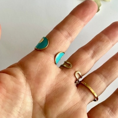 Mão com quatro anéis dourados com detalhes azuis sobre fundo branco com flor amarela.