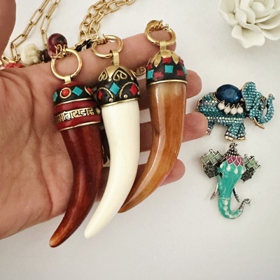 Colares com pendentes em forma de dente coloridos e dois broches de elefante em fundo branco.