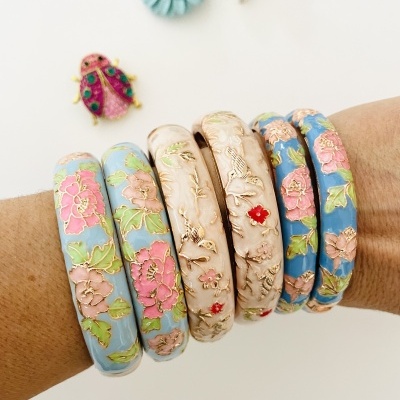 Pulseiras rígidas florais em tons azuis, rosa e dourado num braço com fundo branco e decorativos coloridos